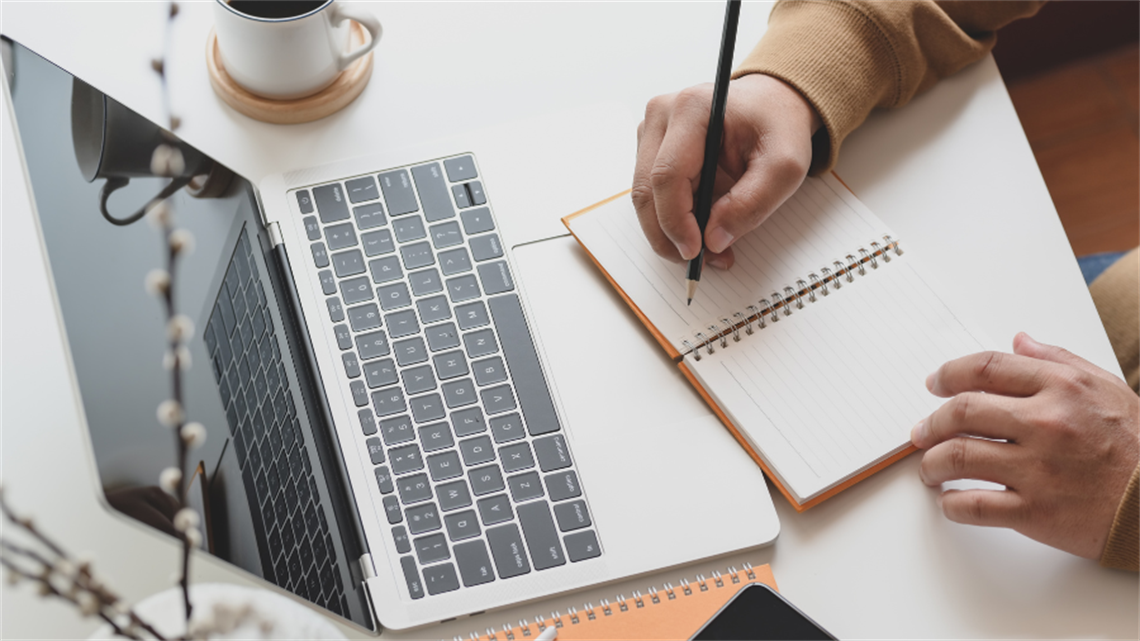 Hand holding pen, writing on notepad with laptop and cup of coffee on desk Hand holding pen, writing on notepad with laptop and cup of coffee on desk